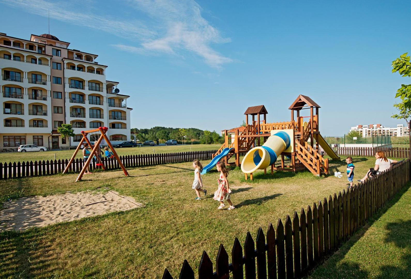 Sunrise All Suites Resort bērnu rotaļu laukums ar slidkalniņiem un šūpolēm uz zālāja pie viesnīcas ēkas saulainā laikā.