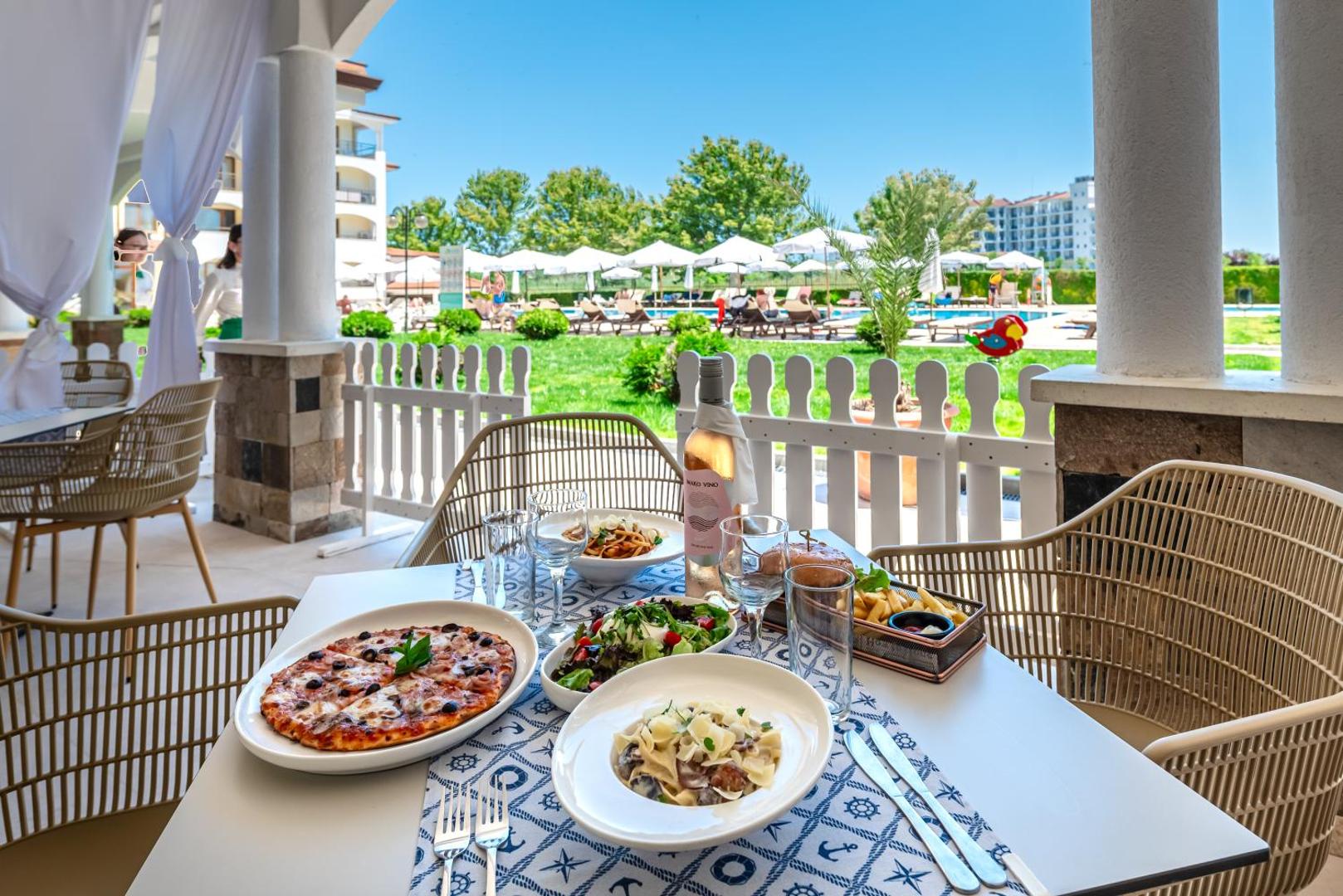 Sunrise All Suites Resort restorāna terase ar skatu uz baseinu, kur uz galda servēta pica, pasta un vīns saulainā dienā.