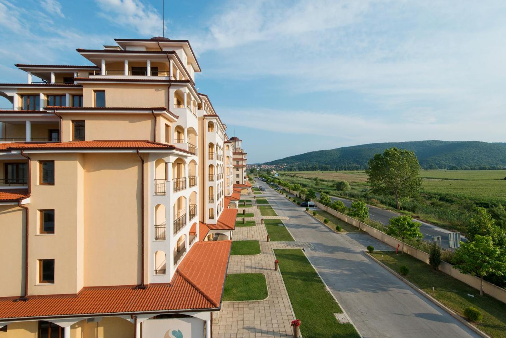 Skats no Sunrise All Suites Resort balkona uz viesnīcas fasādi, piebraucamo ceļu un zaļajiem Bulgārijas kalniem tālumā