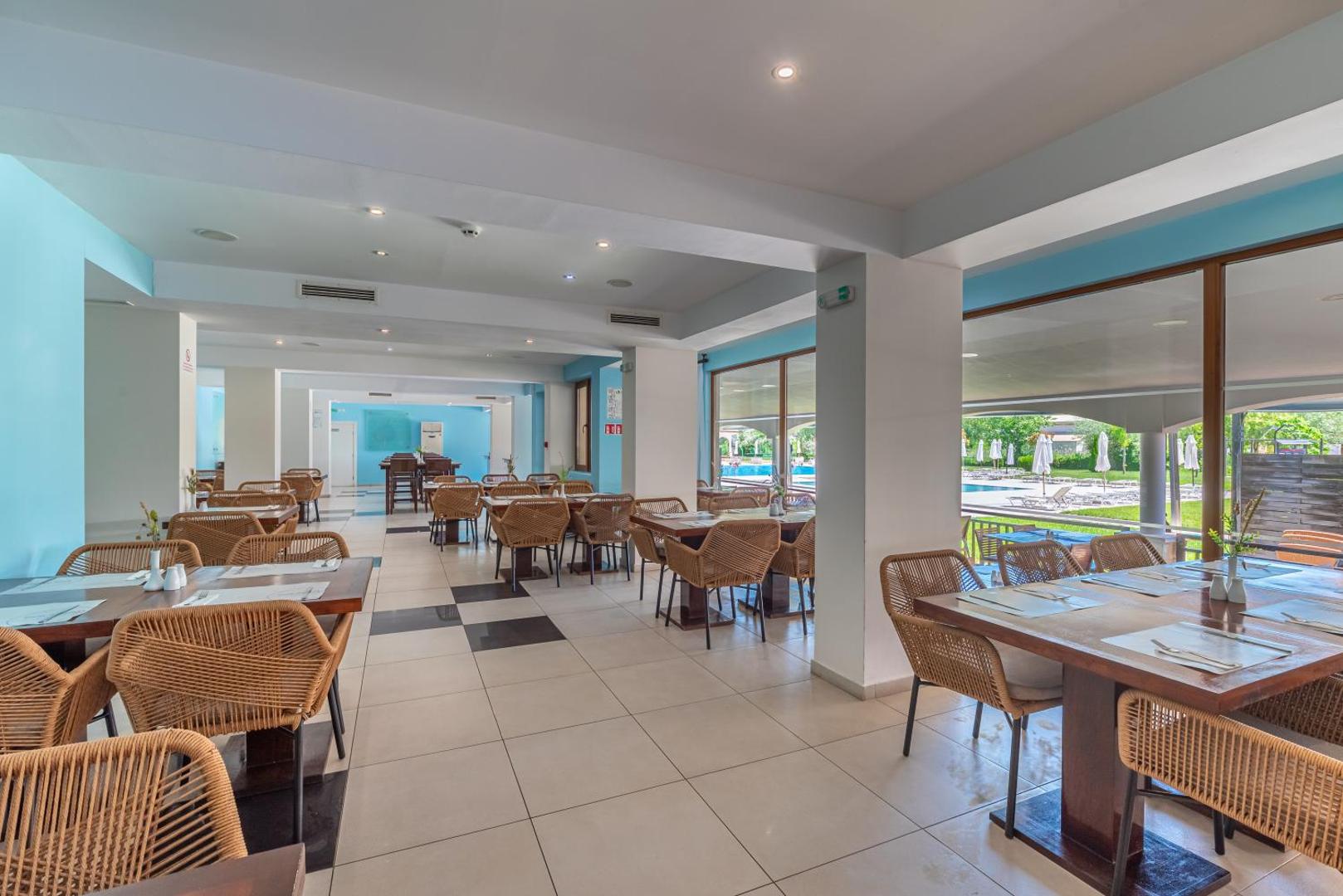 Sunrise All Suites Resort gaišais restorāns ar pītiem krēsliem un skatu caur lielajiem logiem uz viesnīcas baseinu