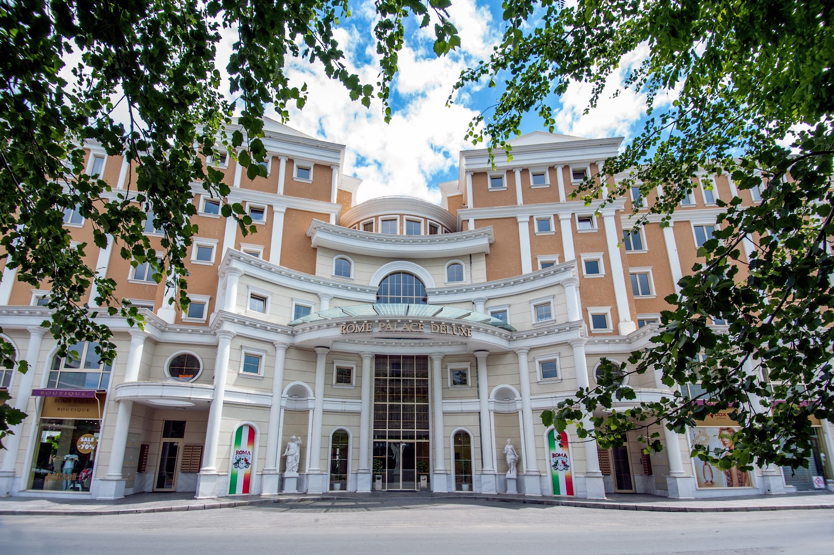 Rome Palace Deluxe centrālā ieeja dienas gaismā caur koku lapotni — klasiskā stila ēka ar kupolu un kolonnām