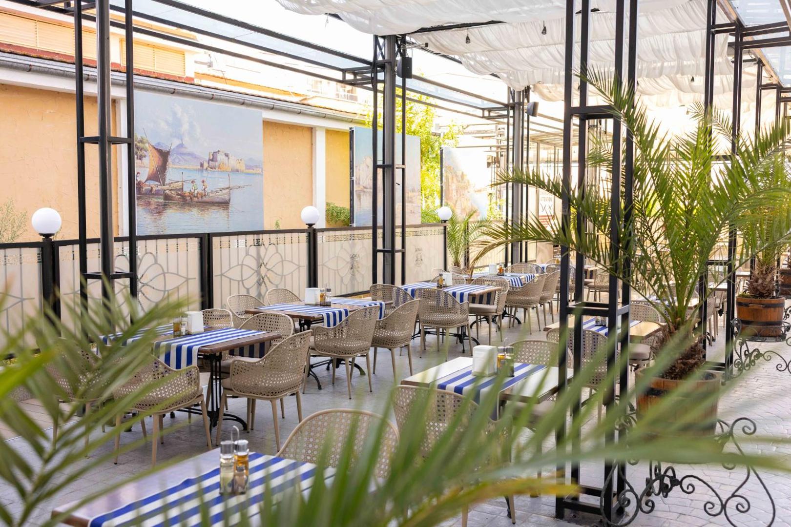 Rome Palace Deluxe restorāna terases skats caur palmu lapām uz galdiņiem ar ziliem galdautiem un mākslas darbiem pie sienām