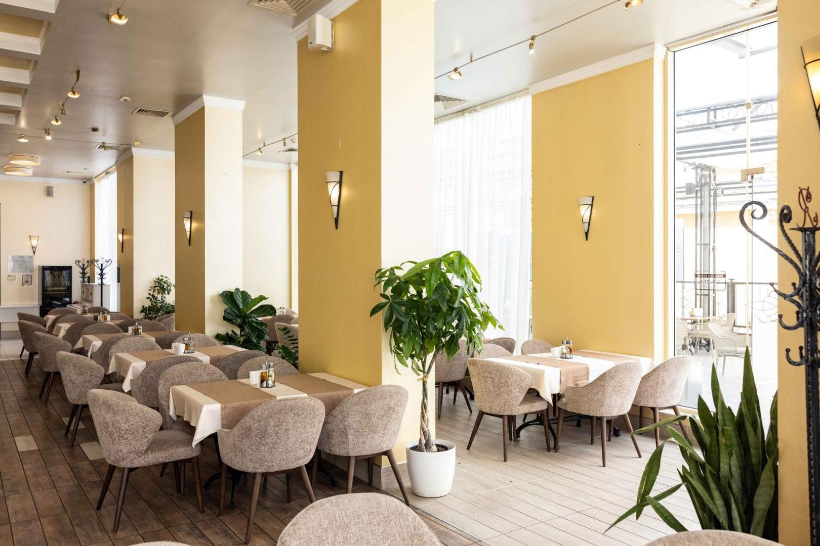 Rome Palace Deluxe iekštelpu restorāna zona ar dzeltenām sienām, mīkstiem krēsliem, lieliem logiem un telpaugiem gaišā atmosfērā