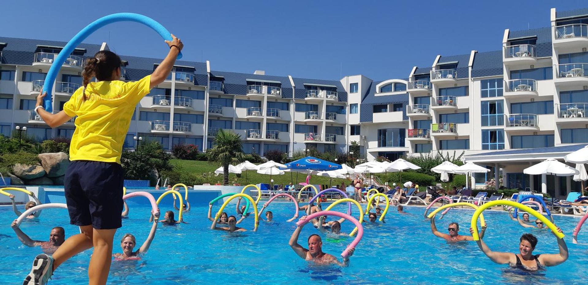 Ūdens aerobikas nodarbība Sineva Beach āra baseinā ar animatoru un viesiem, fonā redzama viesnīcas ēka un saulessargi.