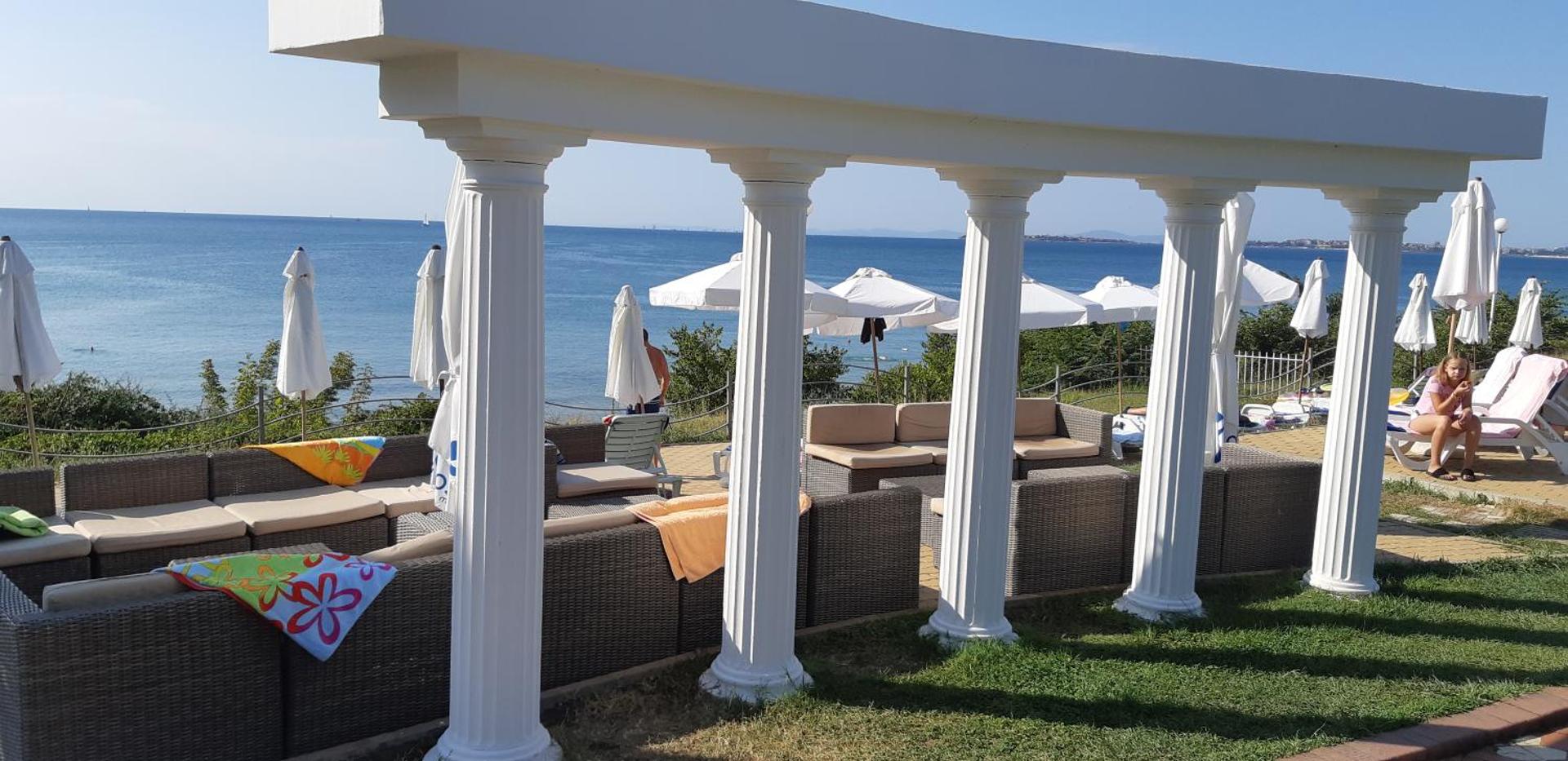 Atpūtas zona ar dīvāniem zem baltām kolonnām Sineva Beach piekrastē ar skatu uz zilo Melno jūru un saulessargiem.