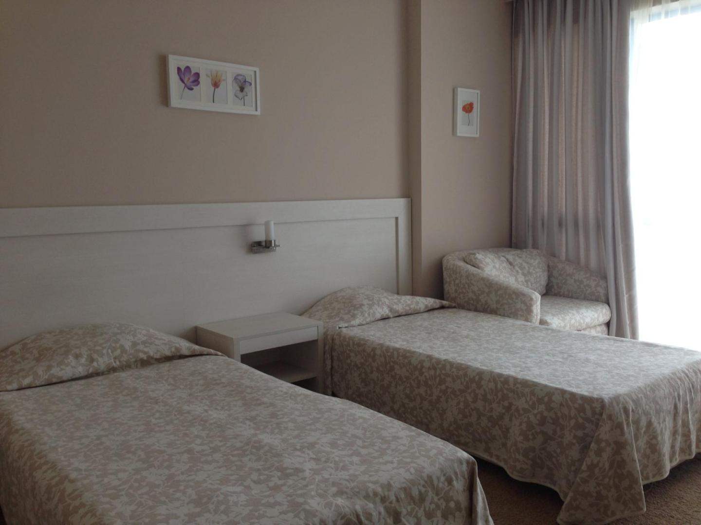 Sineva Beach viesnīcas numurs ar divām gultām, gaišu interjeru un atpūtas krēslu pie loga Sveti Vlas kūrortā Bulgārijā