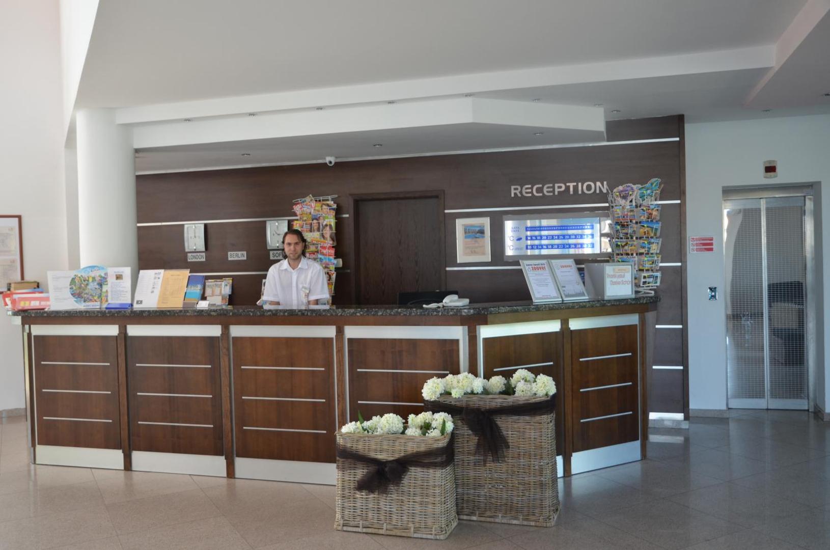 Sineva Beach viesnīcas recepcijas lete ar darbinieku un informatīviem stendiem gaišā un plašā vestibila zonā