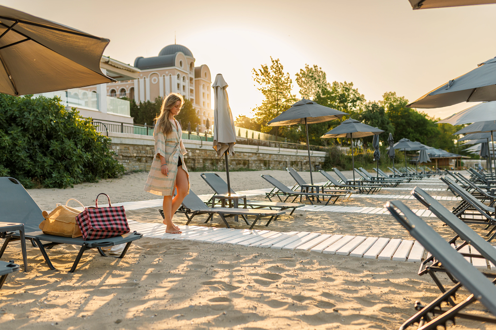 Sieviete saulrietā pastaigājas pa Alua Sunny Beach Resort & Spa pludmali, fonā redzama viesnīcas galvenā ēka