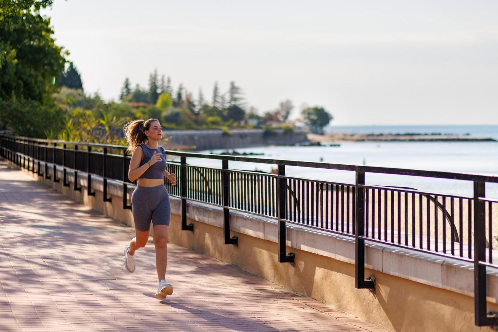 Sieviete skrien pa Alua Sunny Beach Resort & Spa piekrastes promenādi Saulainajā krastā, fonā redzama jūra un koki saulainā dienā.