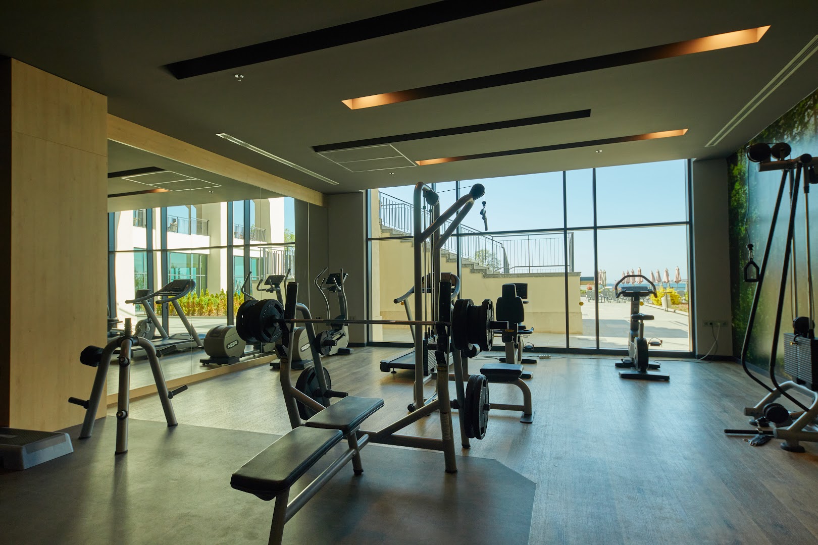Alua Sunny Beach Resort & Spa modernā sporta zāle ar panorāmas logiem, spēka trenažieriem un skatu uz viesnīcas āra teritoriju.
