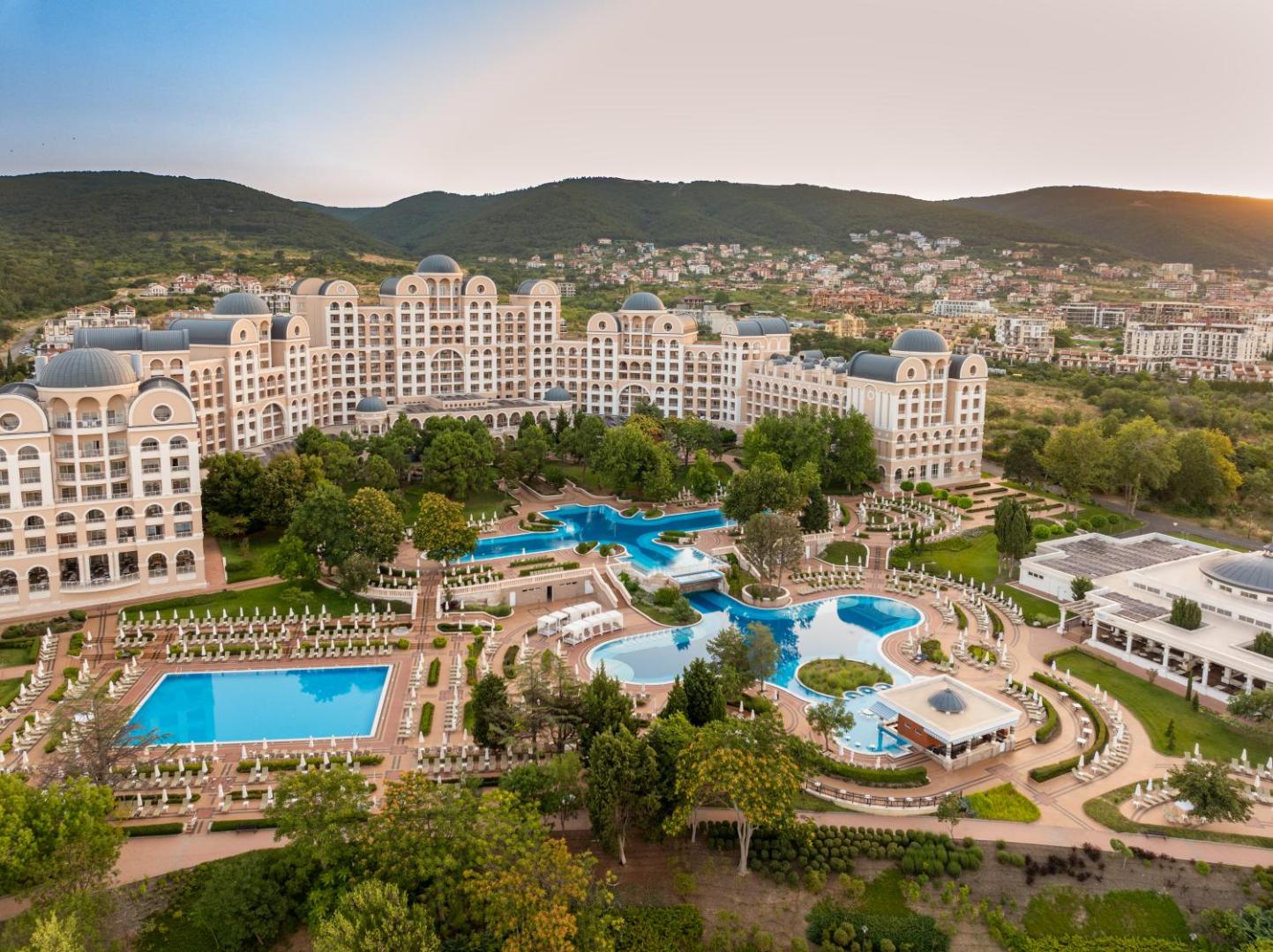 Alua Sunny Beach Resort & Spa arhitektūra un baseinu komplekss saulrietā Bulgārijas piekrastē ar skatu uz kalniem un pilsētu