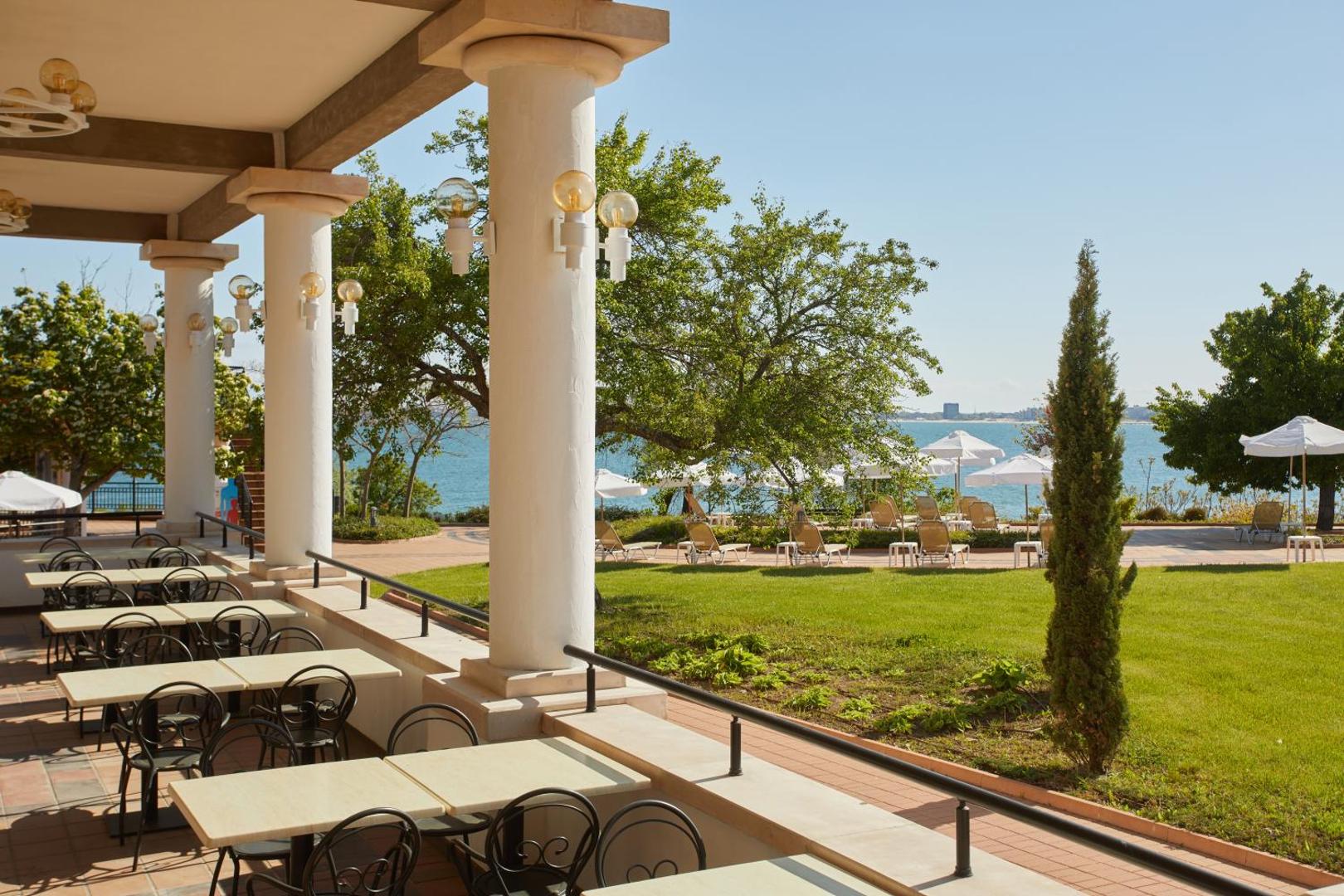 Alua Sunny Beach Resort & Spa āra terase ar galdiņiem, ko ieskauj zaļš zāliens un skats uz jūras piekrasti.