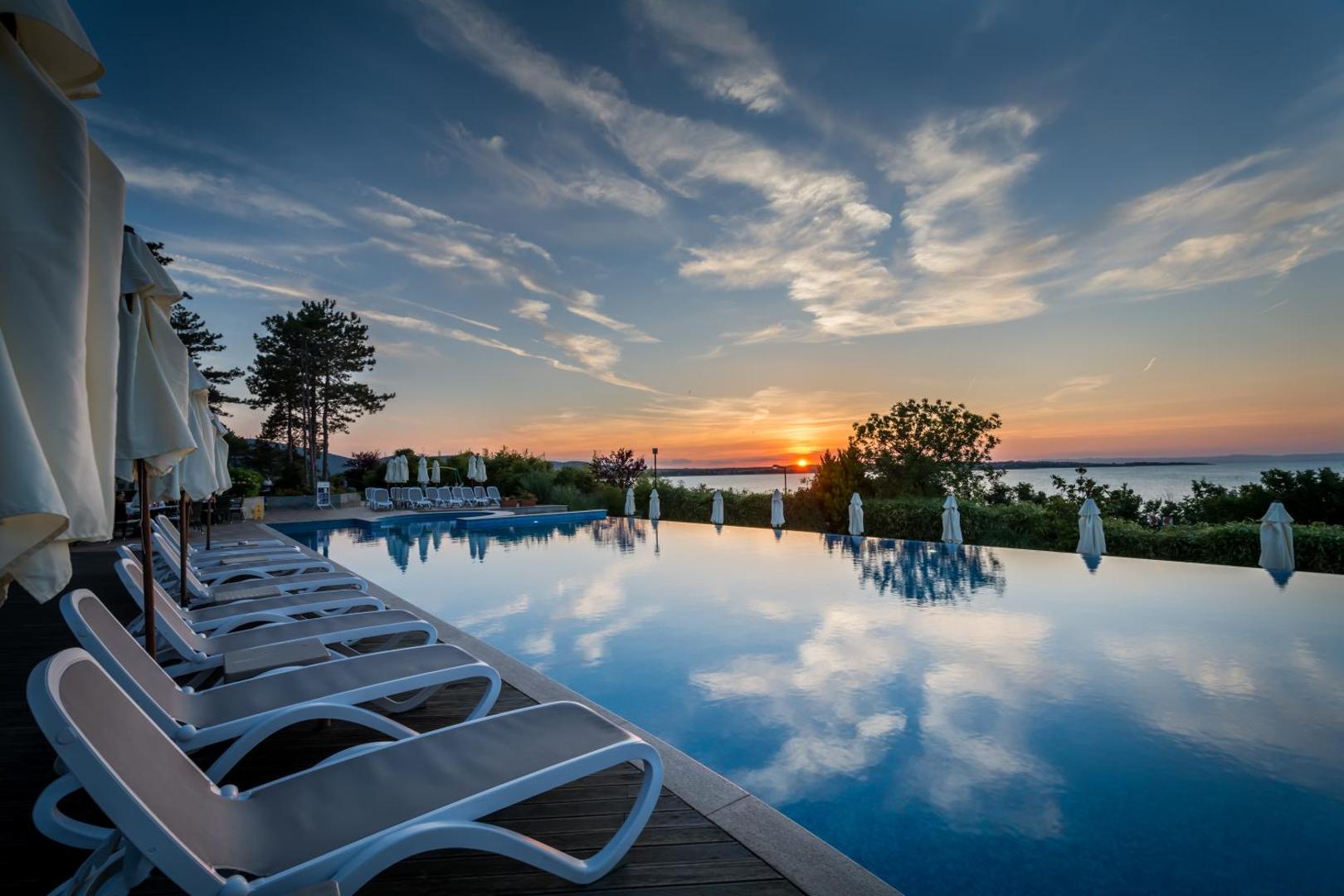 Saulriets pie Viva Mare Beach Hotel bezmalas baseina ar atspulgiem ūdenī un sauļošanās krēsliem piekrastē