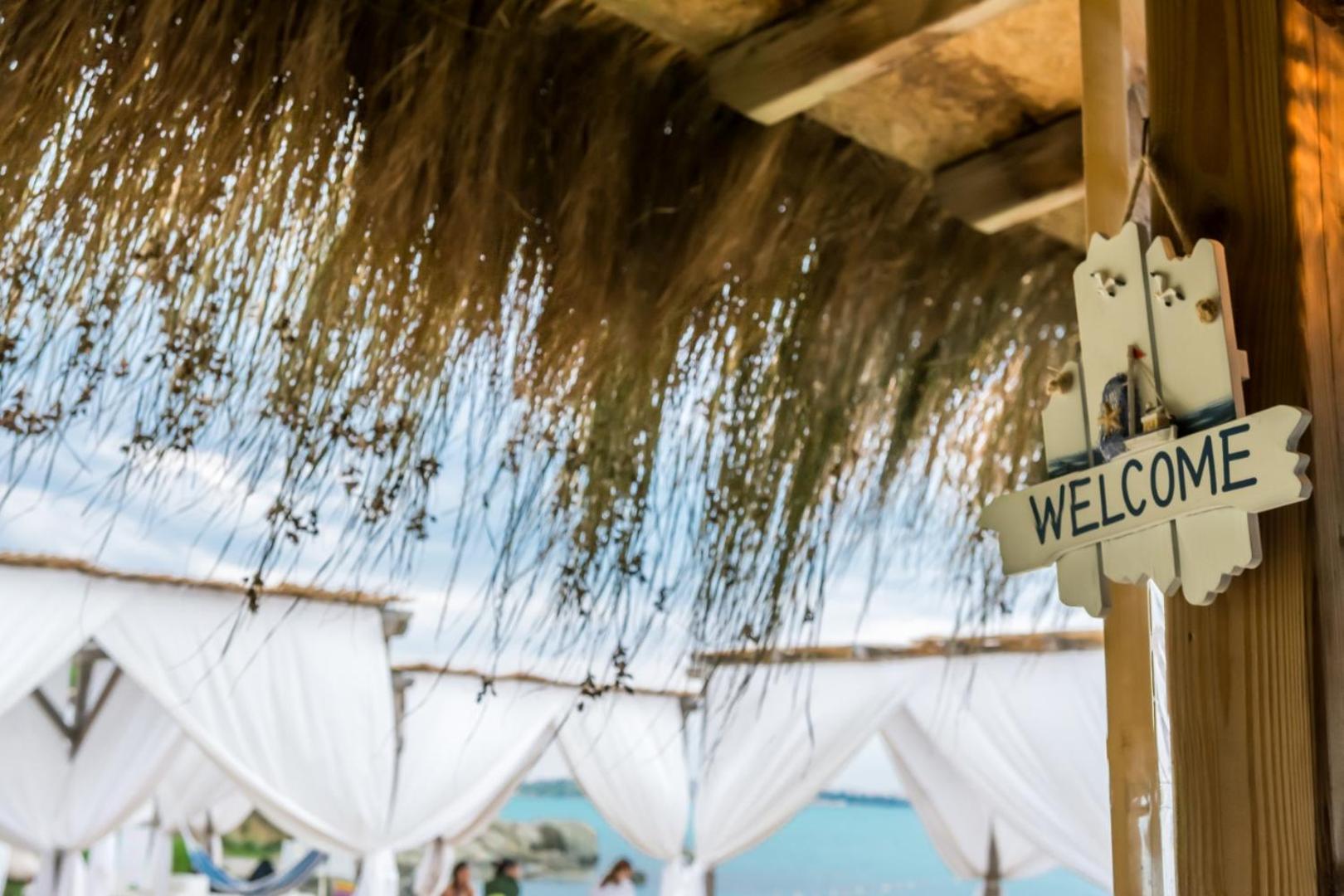 Viva Mare Beach Hotel pludmales zonas dekorācija ar uzrakstu Welcome zem niedru jumta, fonā redzamas baltas atpūtas teltis pie jūras