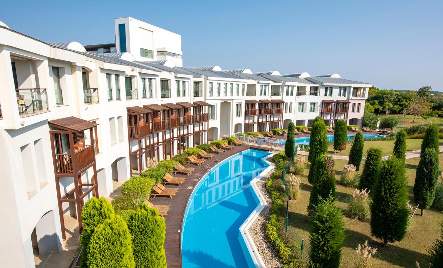 Lykia World Antalya viesnīcas korpuss ar gaišiem numuriem un garu, līkumotu āra baseinu, ko ieskauj kopti dārzi un cipreses