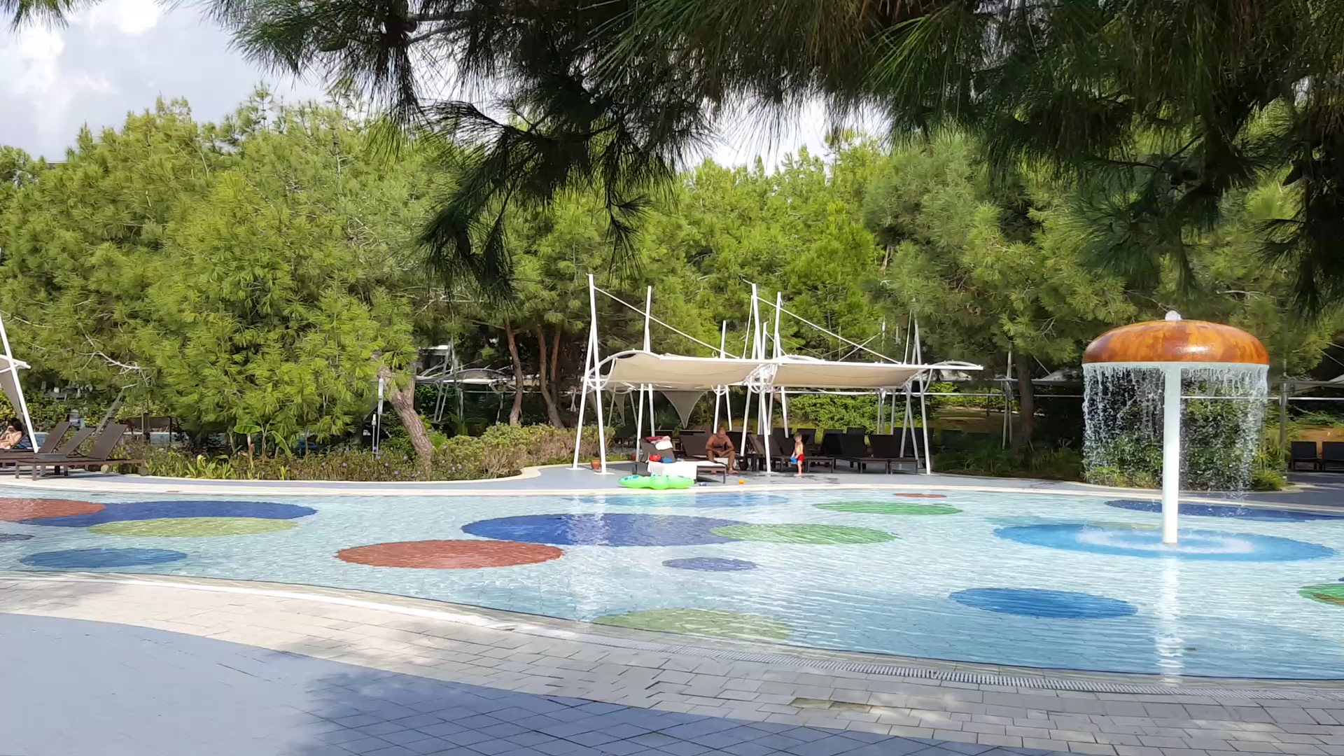 Lykia World Antalya bērnu baseins ar sēnes formas strūklaku, krāsainu pamatni un atpūtas krēsliem priežu meža ielokā