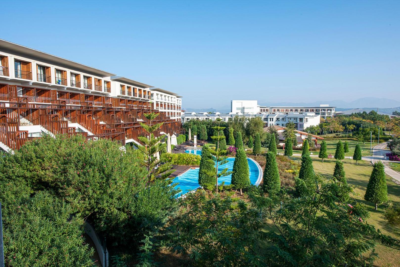 Lykia World Antalya viesnīcas arhitektūra ar koka balkoniem, skatu uz dārzu un baseiniem saulainā dienā Belekas reģionā