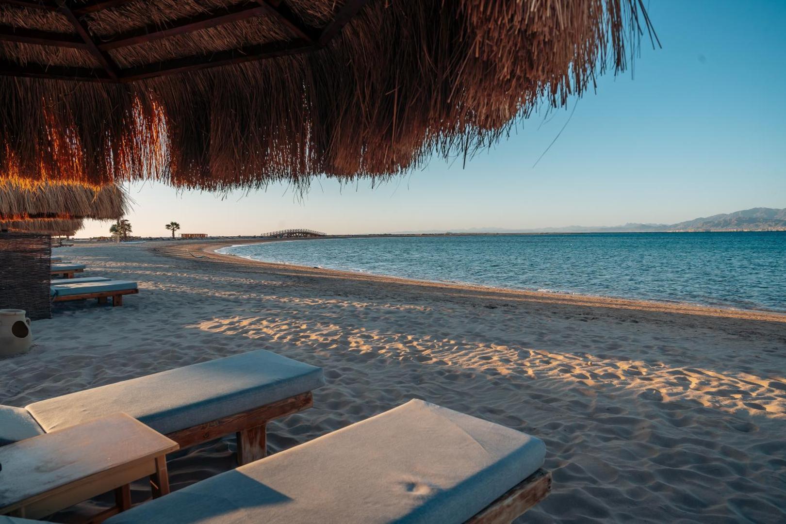 Atpūtas zona ar sauļošanās krēsliem un salmu saulessargiem Sheraton Soma Bay Resort pludmalē — skats uz dzidro jūras līci un kalniem tālumā