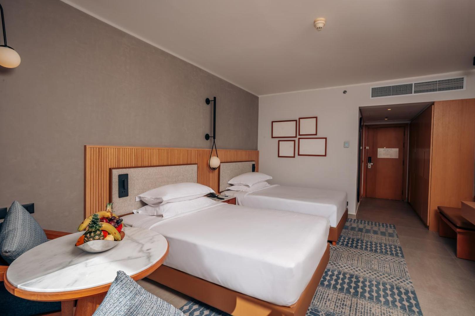 Sheraton Soma Bay Resort numura interjers ar divām gultām, augļu plati uz galdiņa un siltu apgaismojumu