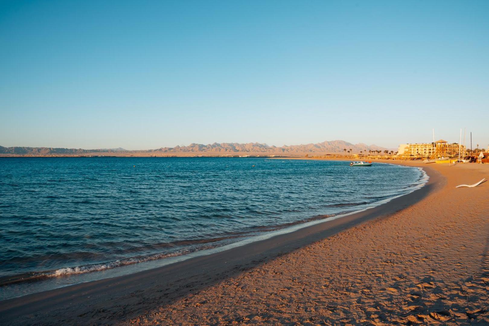 Skats uz Sheraton Soma Bay Resort piekrasti un Sarkanās jūras līci — gaišas smiltis, zils ūdens un kalnu ainava fonā