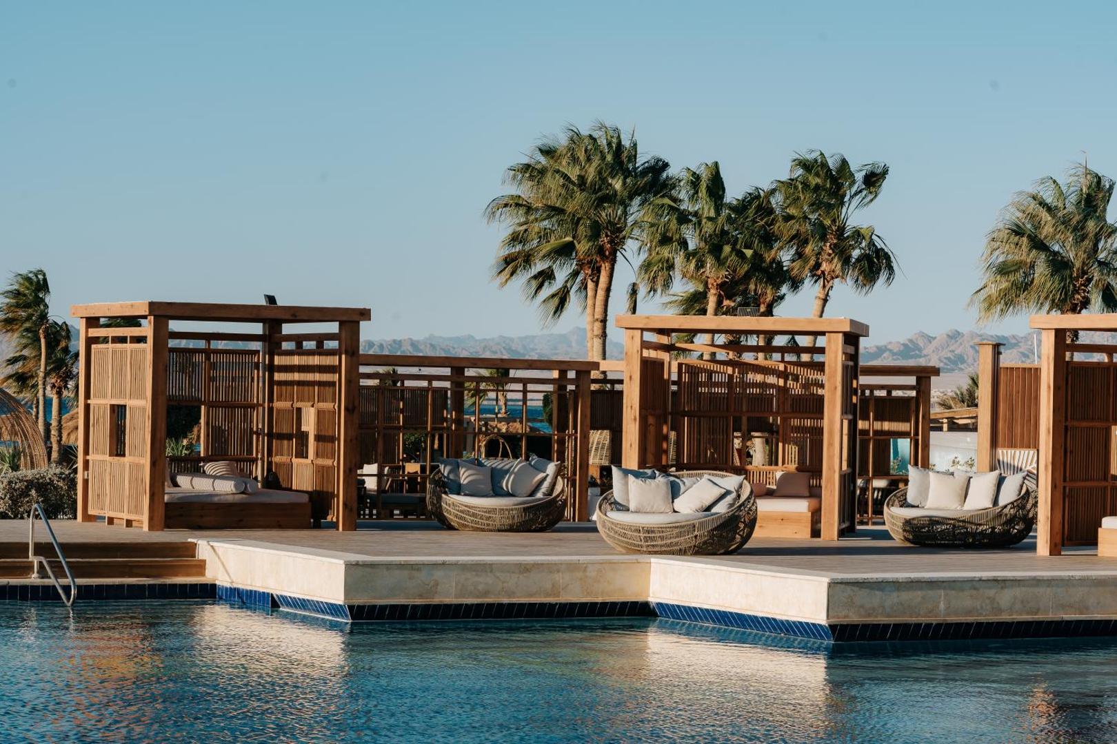 Sheraton Soma Bay Resort atpūtas zona pie baseina ar koka lapenēm, mīkstiem zviļņiem un palmām Hurgadas reģionā, Ēģiptē