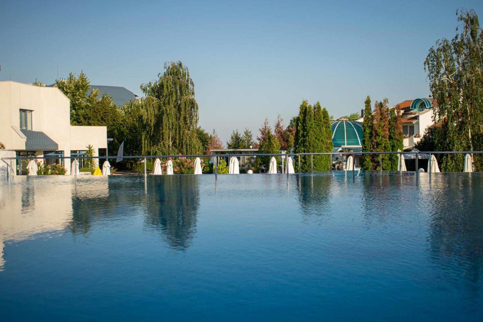 Sineva Park viesnīcas baseins ar pārplūdes malu un skatu uz zaļo teritoriju un kokiem Saulainā krasta kūrortā.