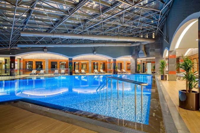 Megasaray Club Belek iekštelpu baseins ar stikla jumta konstrukciju un atpūtas krēsliem — relaksācija spa zonā