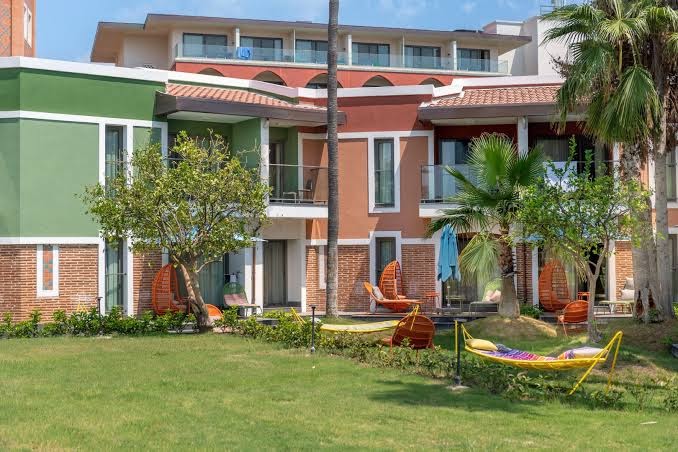 Megasaray Club Belek numuri ar balkoniem un atpūtas zonu dārzā — šūpuļtīkli un krēsli zaļā mauriņā pie viesnīcas korpusa