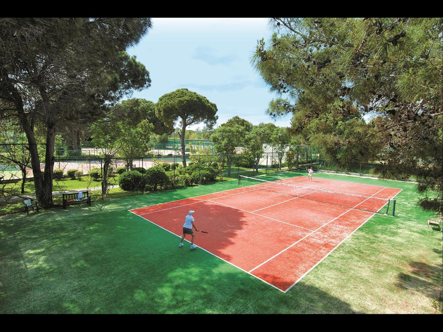 Viesi spēlē tenisu Megasaray Club Belek āra laukumā, ko ieskauj zaļojoši koki un dārzs — sporta aktivitātes kūrorta teritorijā