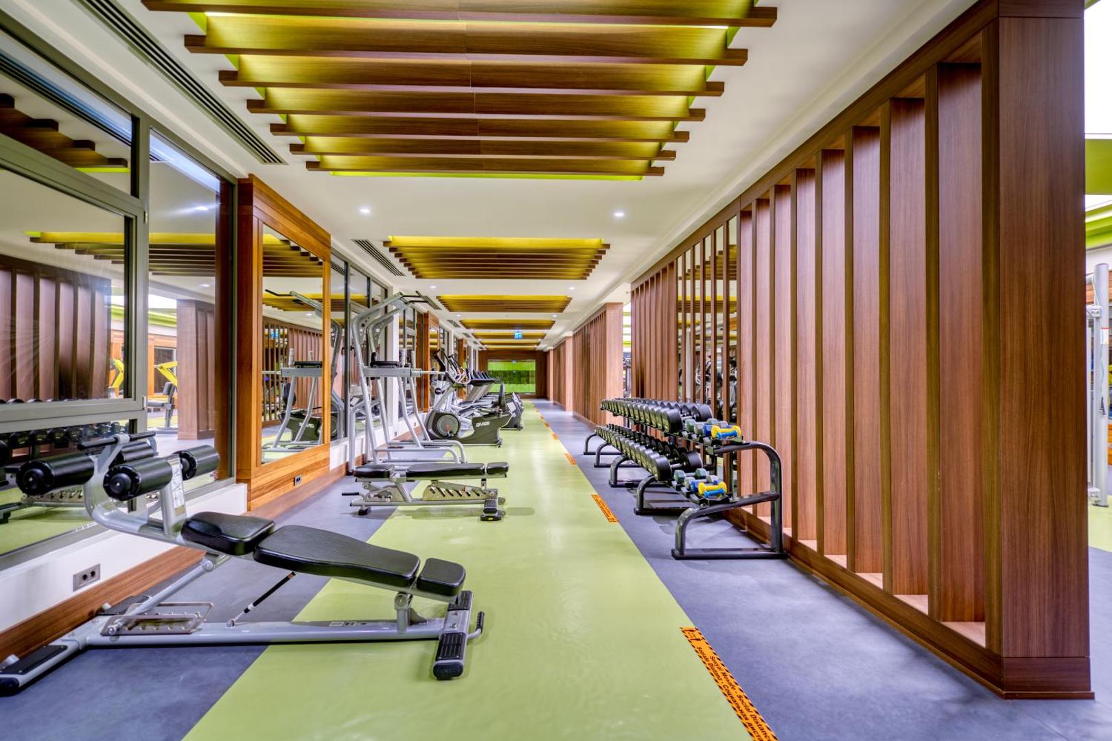 Megasaray Club Belek modernais fitness centrs ar trenažieriem, brīvajiem svariem un koka apdares dizaina elementiem