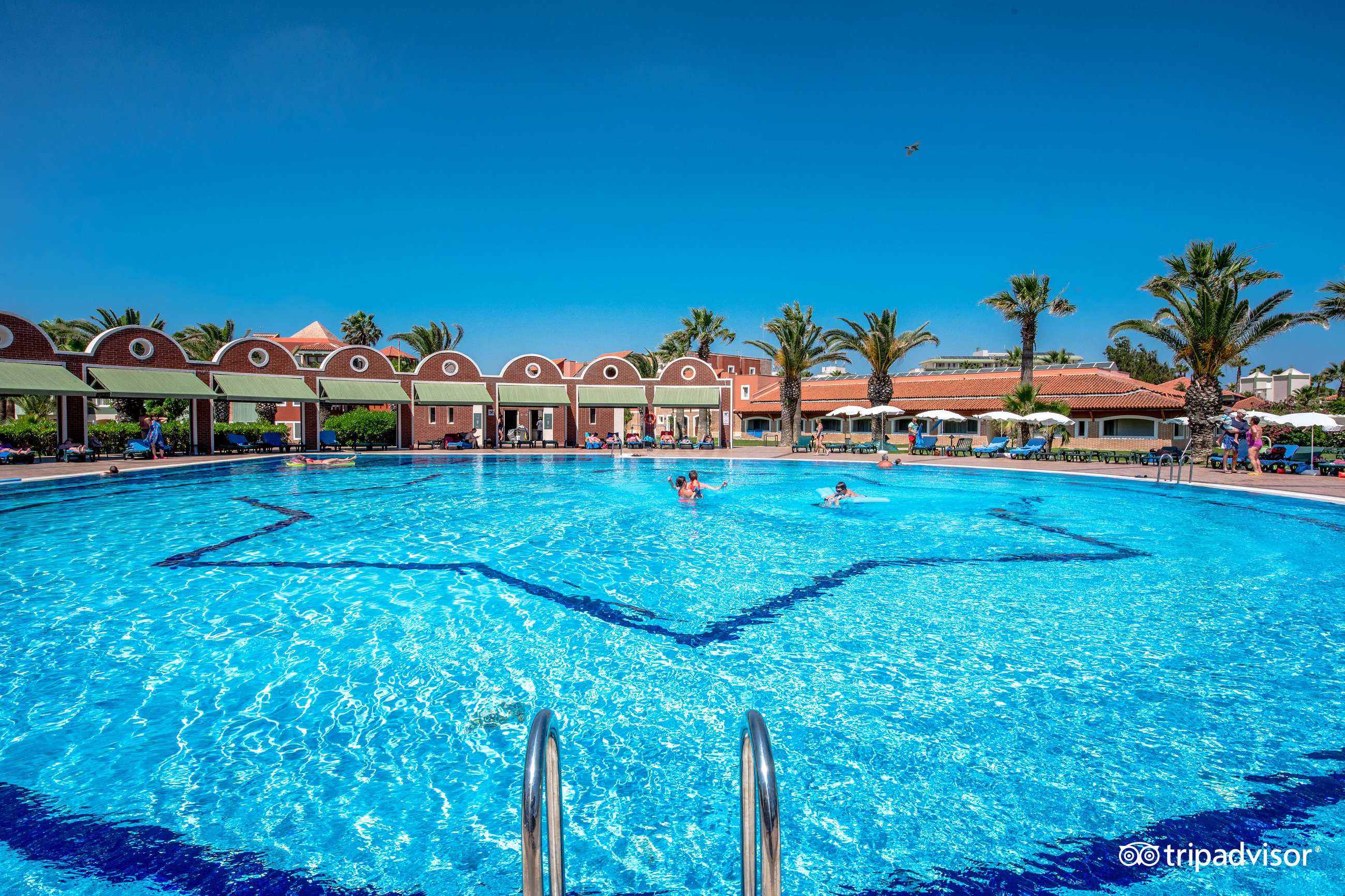 Megasaray Club Belek plašais galvenais āra baseins ar zilu mozaīku rakstu, ko ieskauj palmas un atpūtas krēsli saulainā dienā
