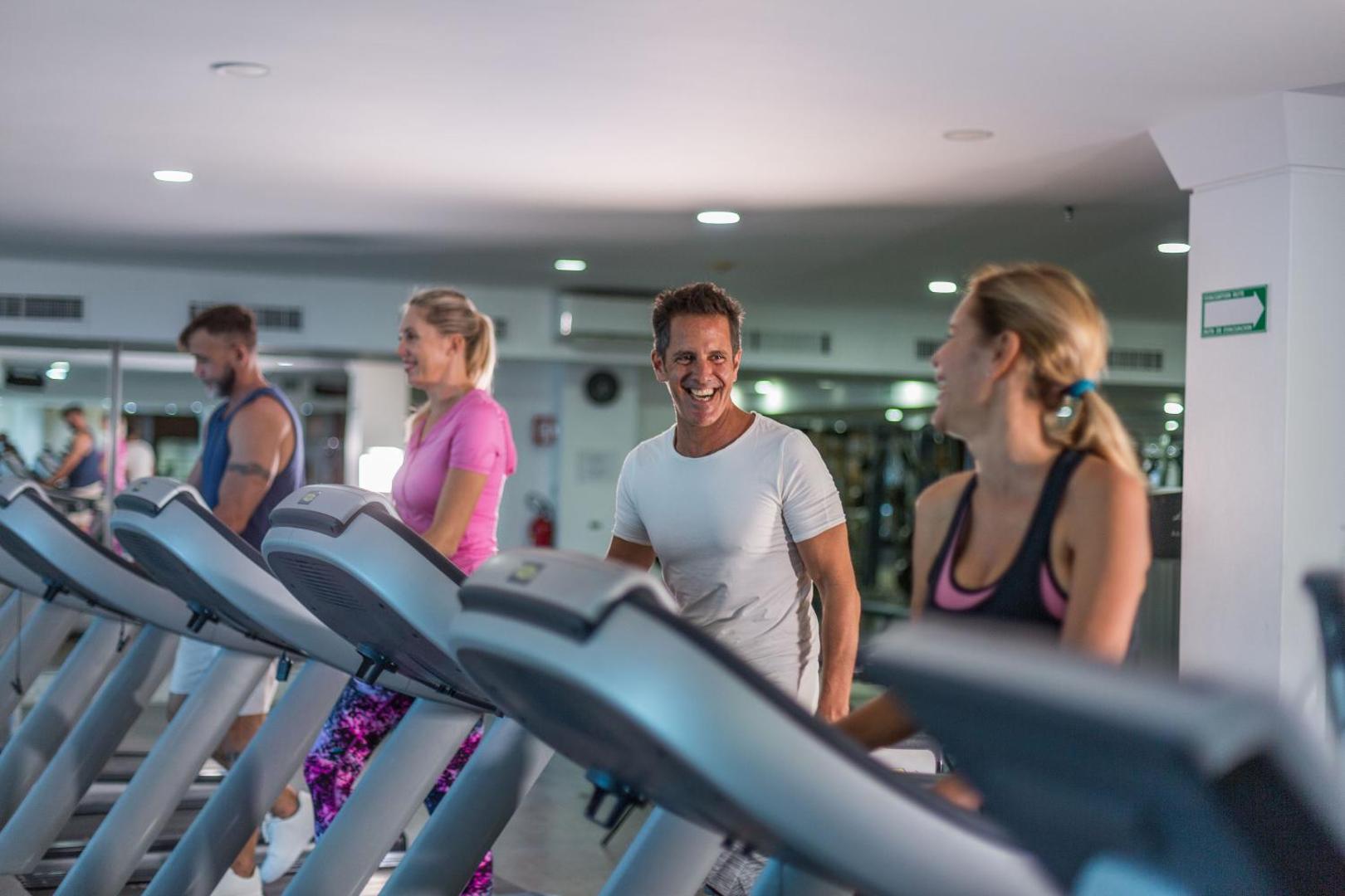 Viesi sporto Sandos Cancun fitnesa centrā uz skrejceļiem — gaiša un moderna trenažieru zāle ar skatu uz Kankunas kūrorta teritoriju.