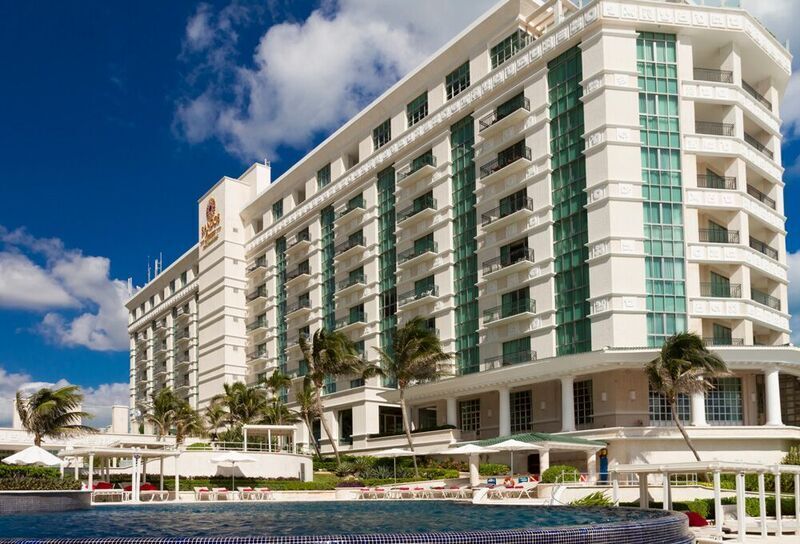 Sandos Cancun viesnīcas galvenā ēka un baseins saulainā dienā ar palmām un atpūtas krēsliem Kankunas kūrortā