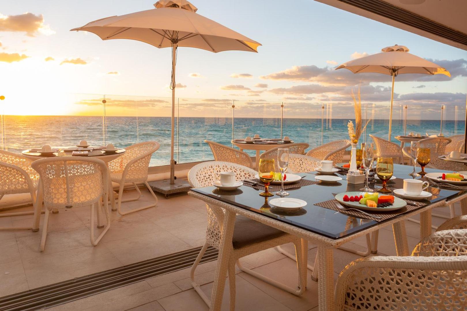 Viesnīcas Sandos Cancun āra terases restorāns saulrieta laikā ar skatu uz okeānu — eleganti klāti galdi un balti saulessargi.