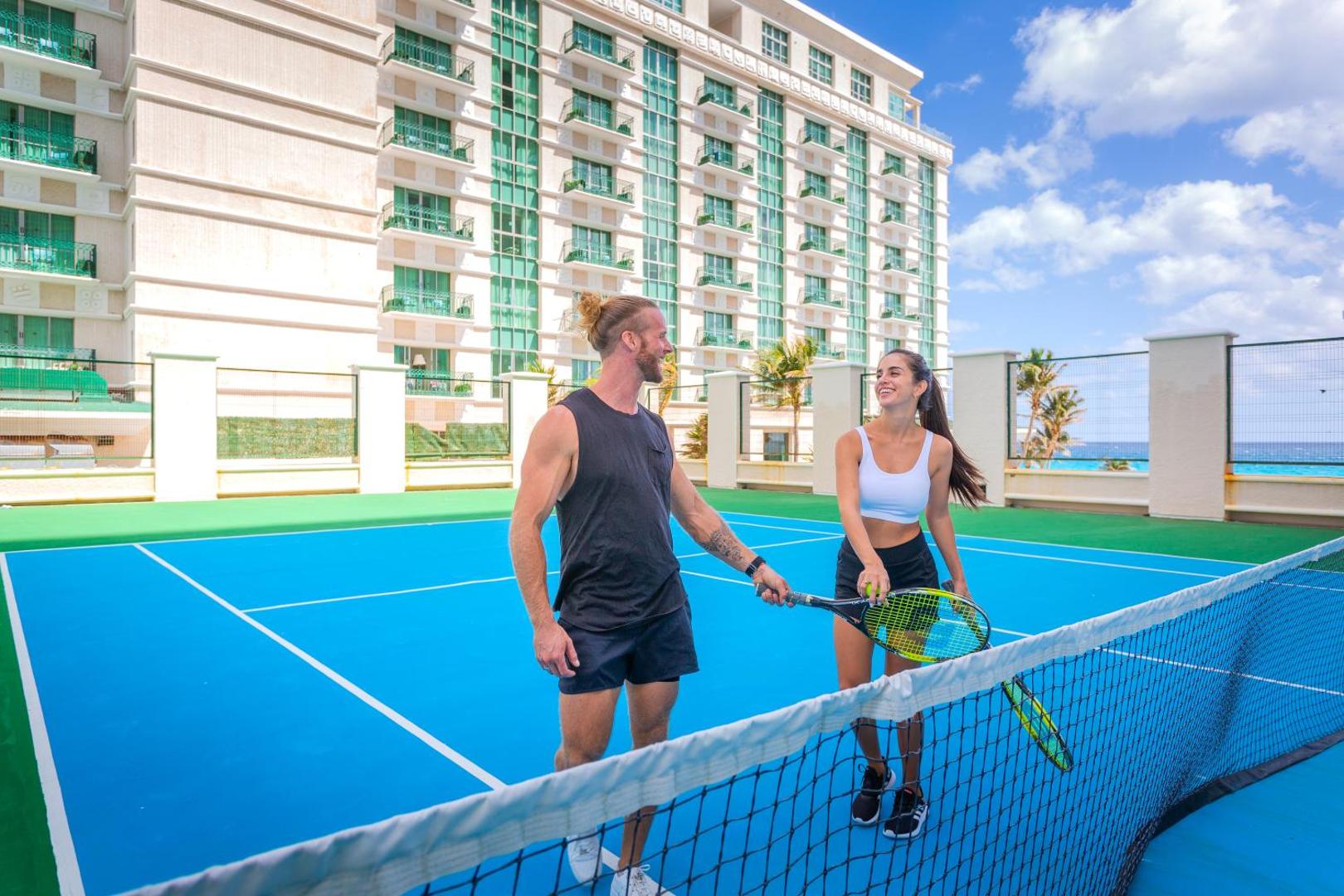 Sandos Cancun tenisa laukums ar zilu segumu pie viesnīcas ēkas Kankunas piekrastē — pāris ar raketēm gatavojas spēlei saulainā dienā.