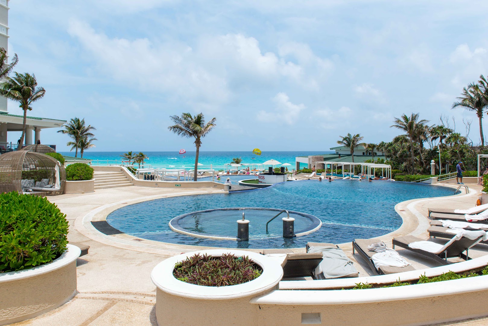 Sandos Cancun kūrorta teritorija ar vairākiem baseiniem, sauļošanās krēsliem un palmām Kankunas piekrastē, fonā tirkīzzila jūra