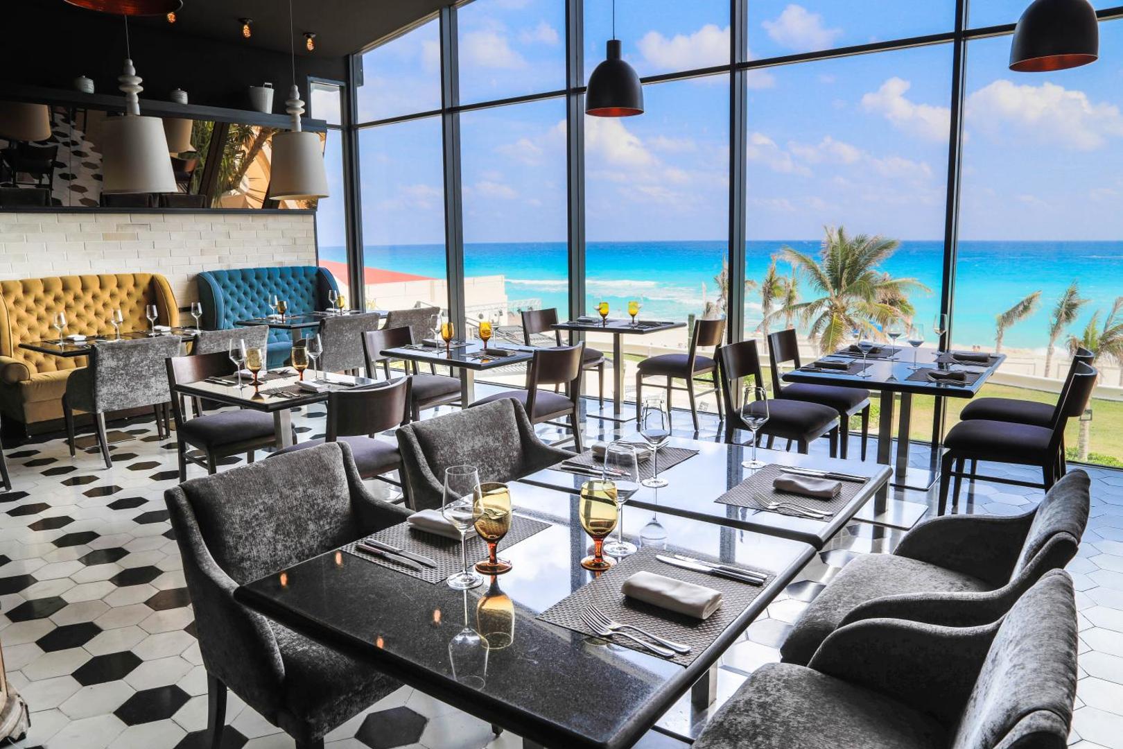 Sandos Cancun restorāna interjers ar panorāmas logiem uz tirkīzzilo jūru — mūsdienīgs dizains, ērti krēsli un palmu skats.