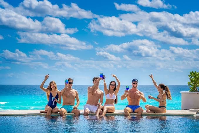 Jaunieši bauda kokteiļus Sandos Cancun baseinā ar pārplūdes malu uz Karību jūras un zilu debesu fona