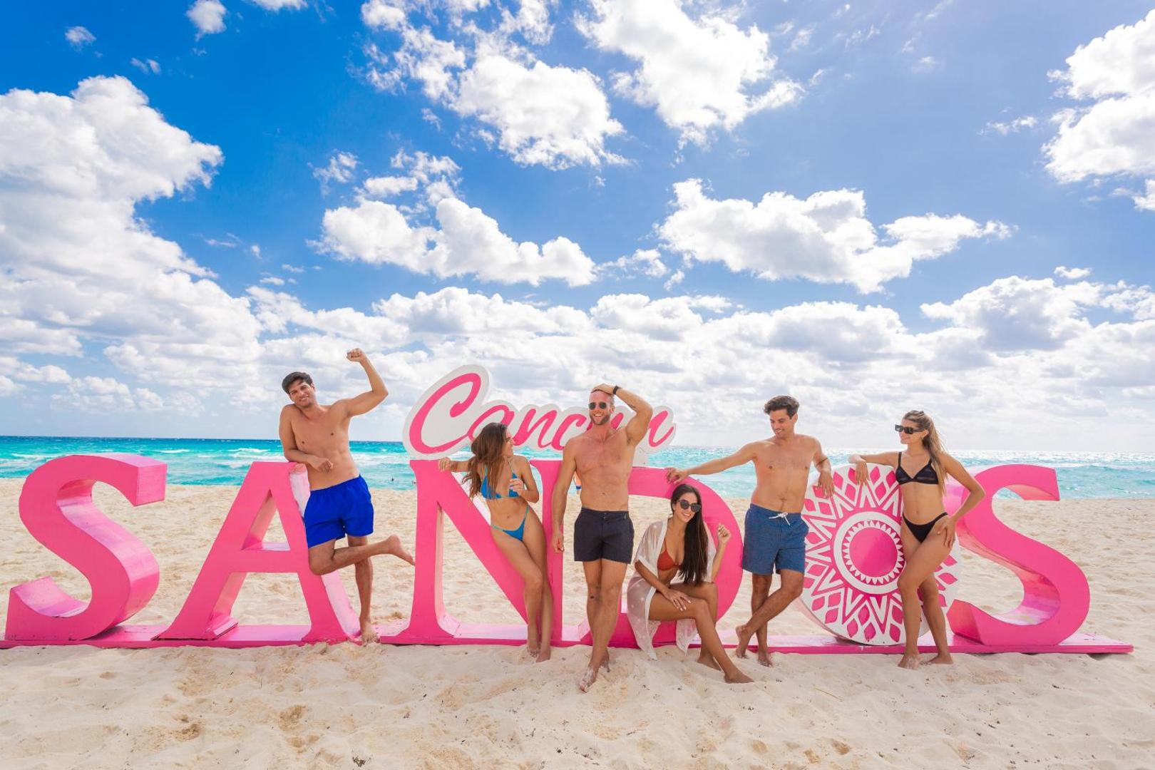Sandos Cancun viesnīcas viesi pozē pie lieliem rozā burtiem Kankūnas pludmales smiltīs — saulains laiks, zila jūra un balti mākoņi.