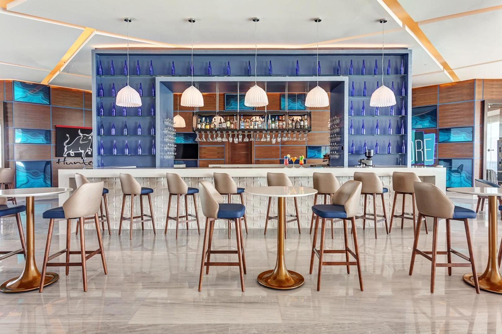 Royalton CHIC Cancun moderns bārs ar zilu fonu, augstiem krēsliem un plašu dzērienu izvēli Kankunā