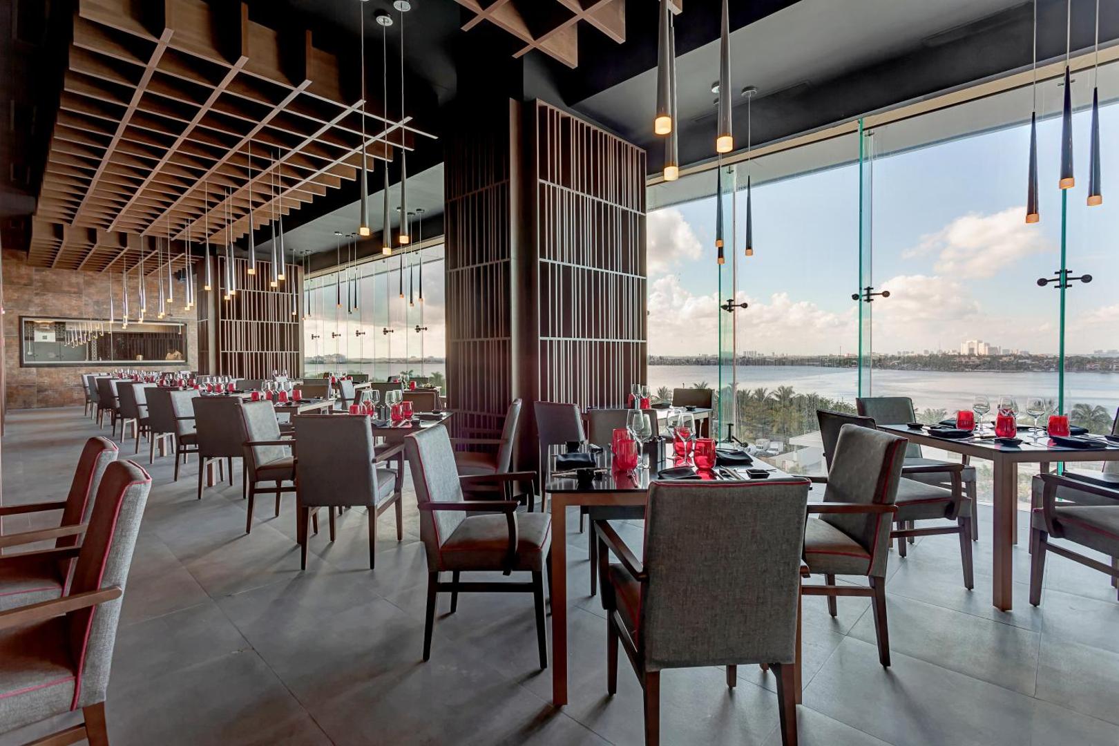 Royalton CHIC Cancun moderns restorāns ar panorāmas logiem, pelēkiem krēsliem un skatu uz lagūnu saulrieta laikā
