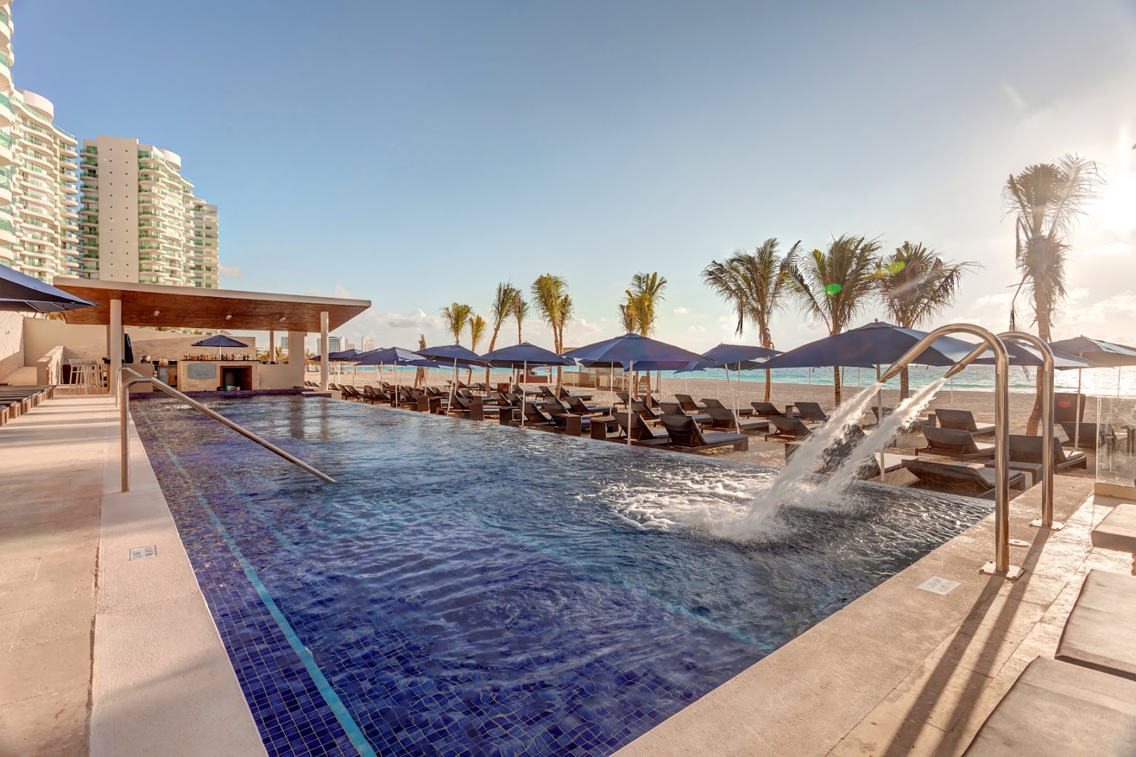 Royalton CHIC Cancun baseins ar masāžas strūklām un skatu uz pludmali, fonā redzama kūrorta modernā arhitektūra