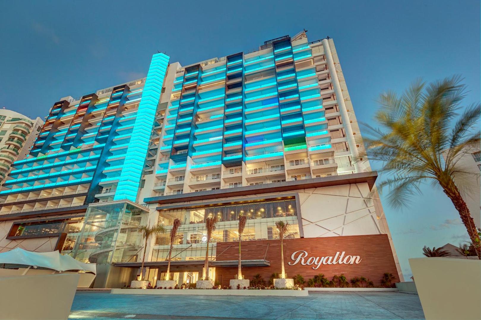 Royalton CHIC Cancun viesnīcas fasāde vakarā ar krāsainu apgaismojumu Kankunā, Meksikā — moderna arhitektūra piekrastē ar palmām un zīmola logotipu