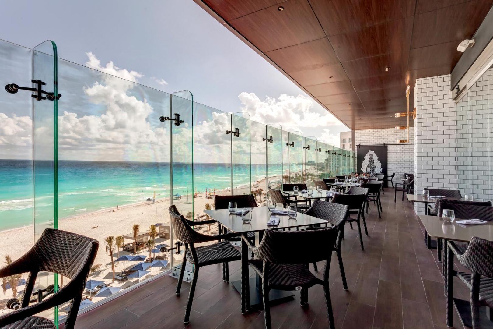 Royalton CHIC Cancun āra terases restorāns ar stikla margām, galdiņiem un skatu uz pludmali un okeānu saulainā dienā