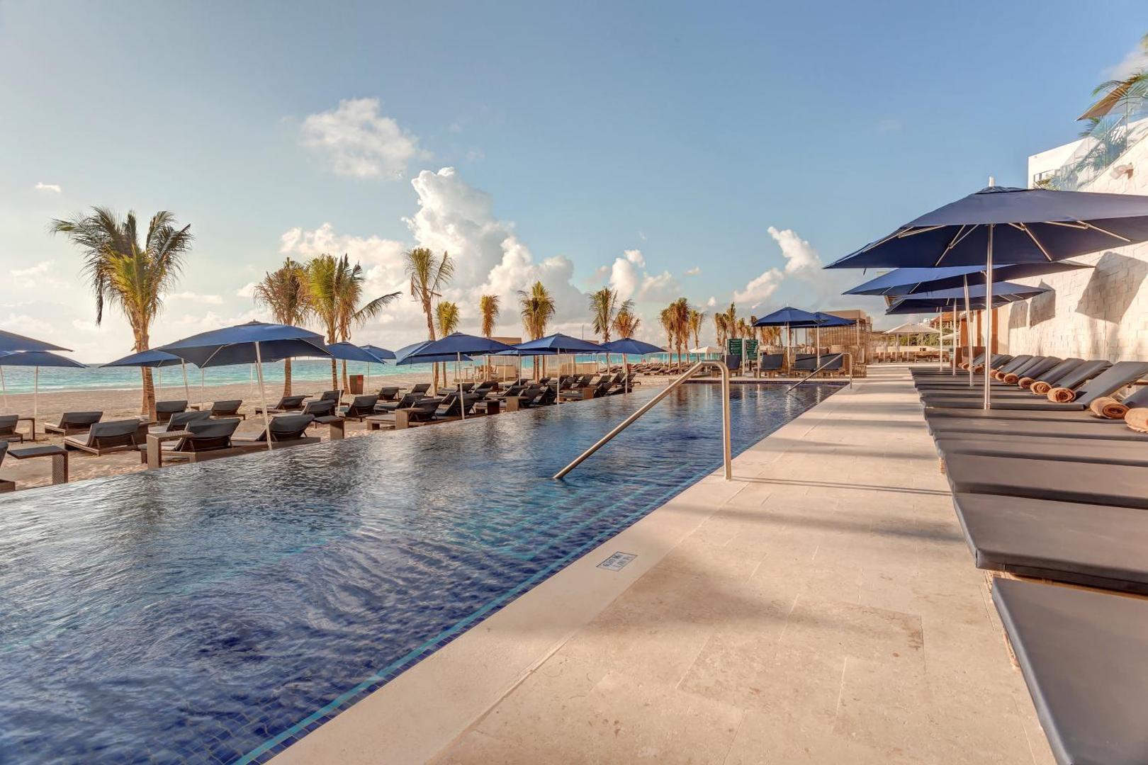 Royalton CHIC Cancun piekrastes baseins ar ziliem saulessargiem un skatu uz balto smilšu pludmali un palmām
