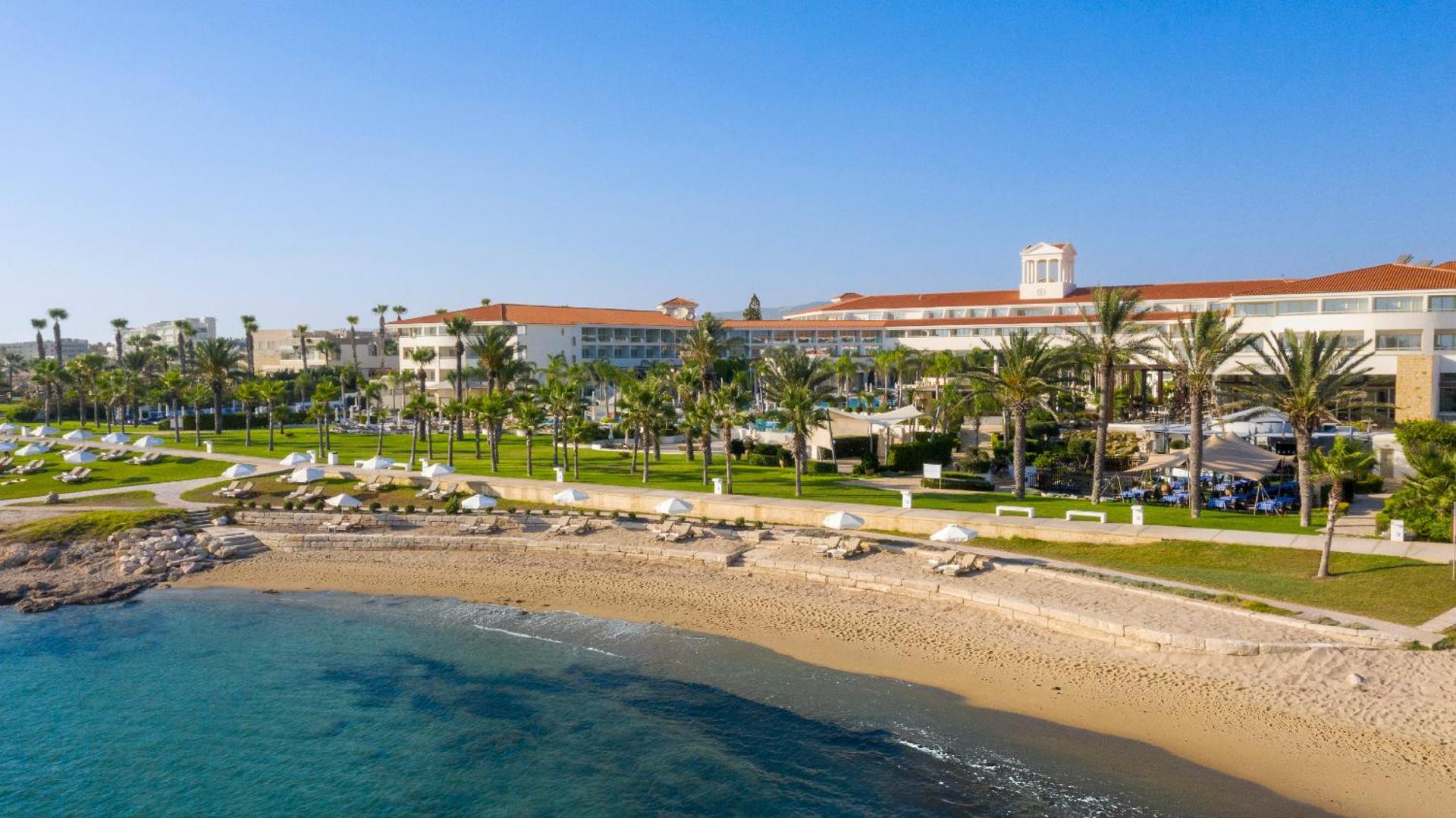 Olympic Lagoon Resort Paphos viesnīcas ēka un labiekārtota smilšu pludmale ar saulessargiem Pafas piekrastē, Kiprā