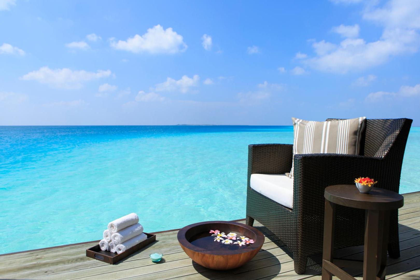 Velassaru Maldives kūrorta terase ar skatu uz tirkīza okeānu, pīts atpūtas krēsls, dvieļi un dekoratīvs trauks ar ziediem