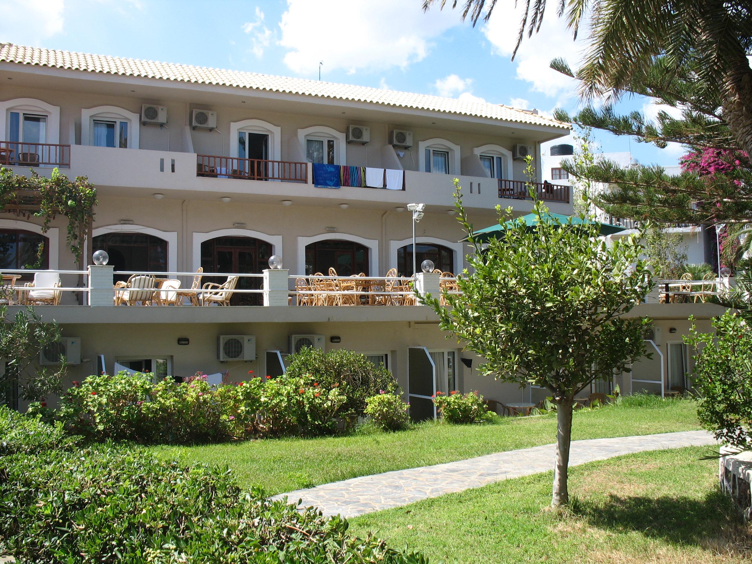 Zeus Palm Beach Hotel trīsstāvu ēka ar balkoniem, zaļu zālienu un koptu dārzu Krētas piekrastē saulainā dienā
