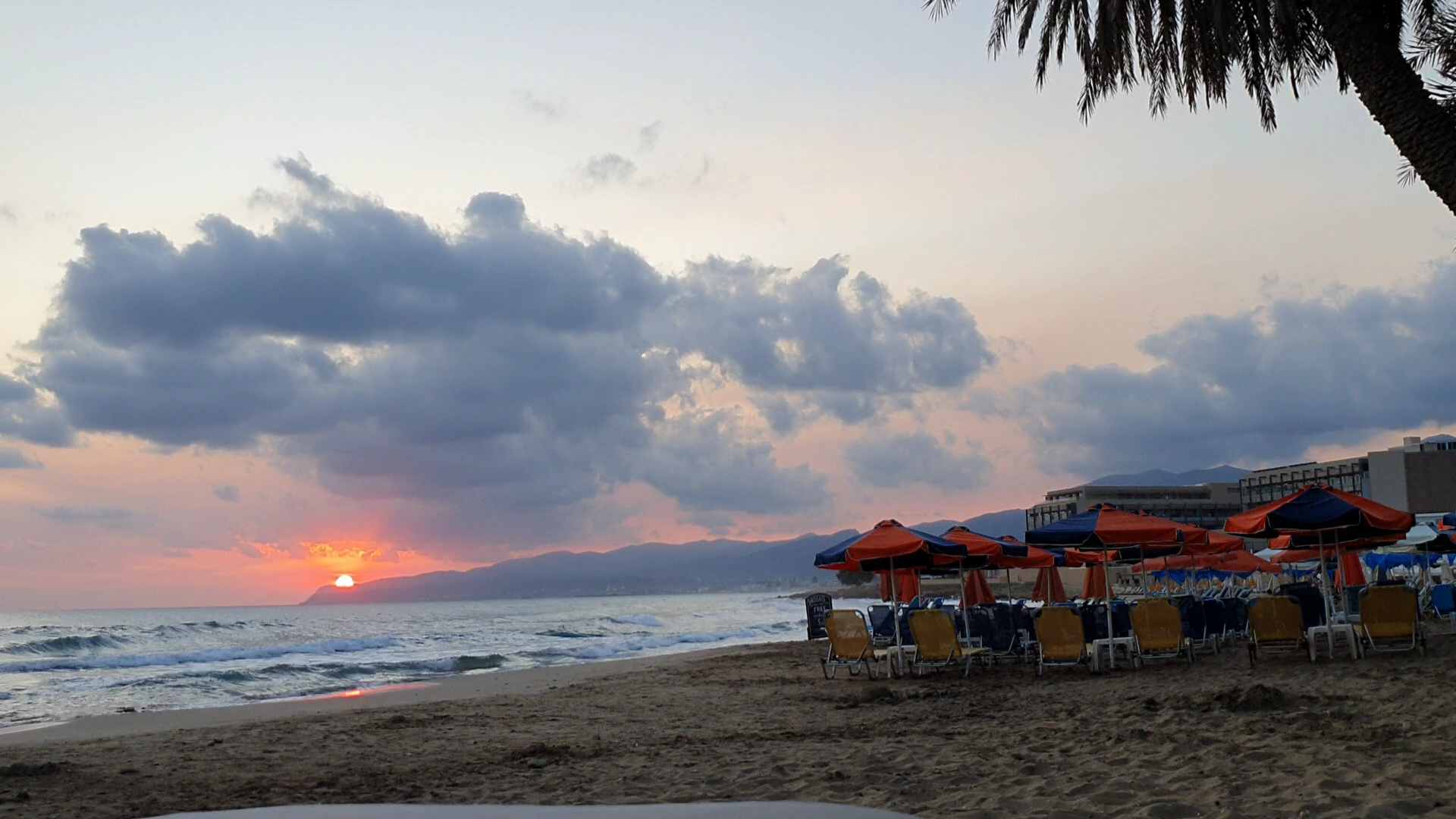 Saulriets Zeus Palm Beach Hotel pludmalē Krētā — skats uz jūru, kalniem un pludmales saulessargiem vakara saules gaismā