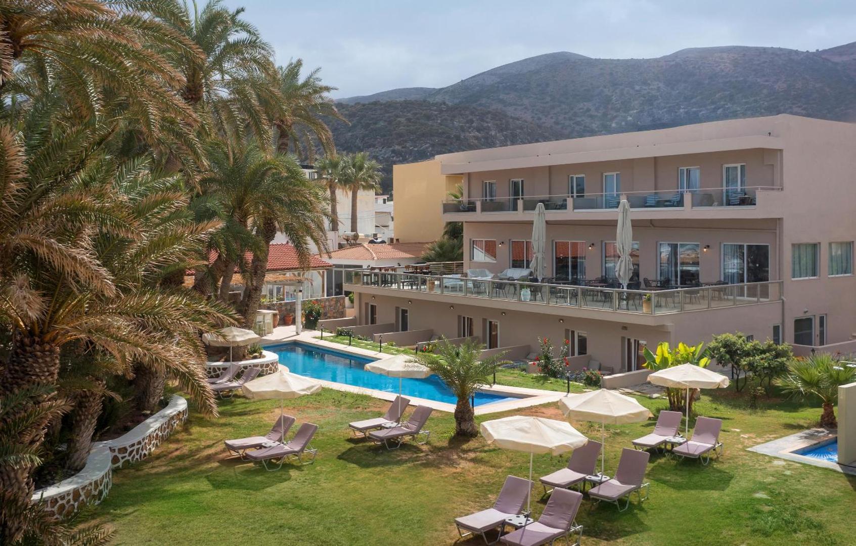 Skats uz Zeus Palm Beach Hotel ēku, baseinu un zaļo dārzu ar atpūtas krēsliem un saulessargiem uz kalnu fona.
